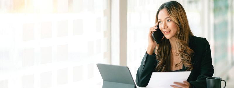 a woman is sitting at a desk with a laptop and talking on a cell phone while holding a piece of paper.