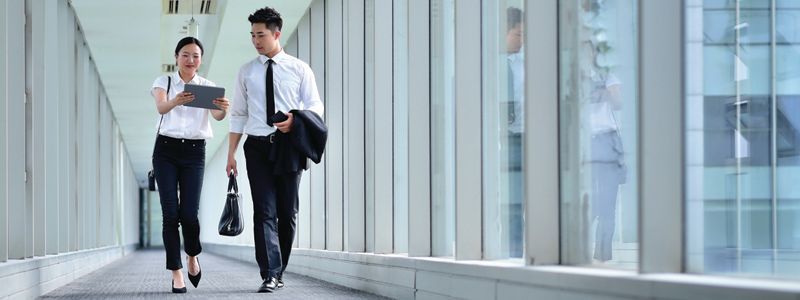 Two office workers walk through a bright glass hallway, talking and carrying bags and papers.