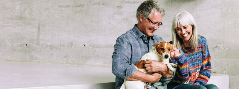 A couple smiles while holding a dog. They sit on a step in front of a concrete wall.