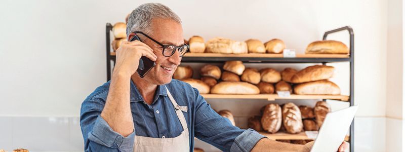 A man is talking on a cell phone in a bakery while looking at a tablet.