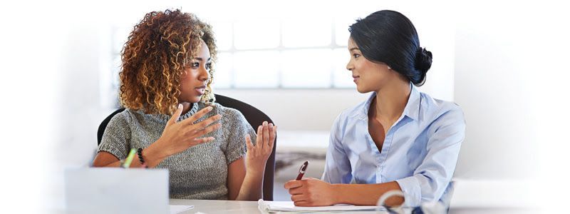 Two women in a well-lit office setting engaged in a discussion. One woman is speaking animatedly, gesturing with her hands, while the other listens attentively, holding a pen and notepad.