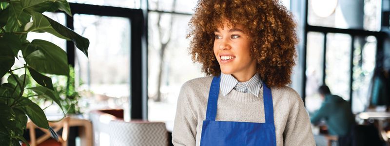 Smiling person in a blue apron standing in a bright café kitchen near large windows