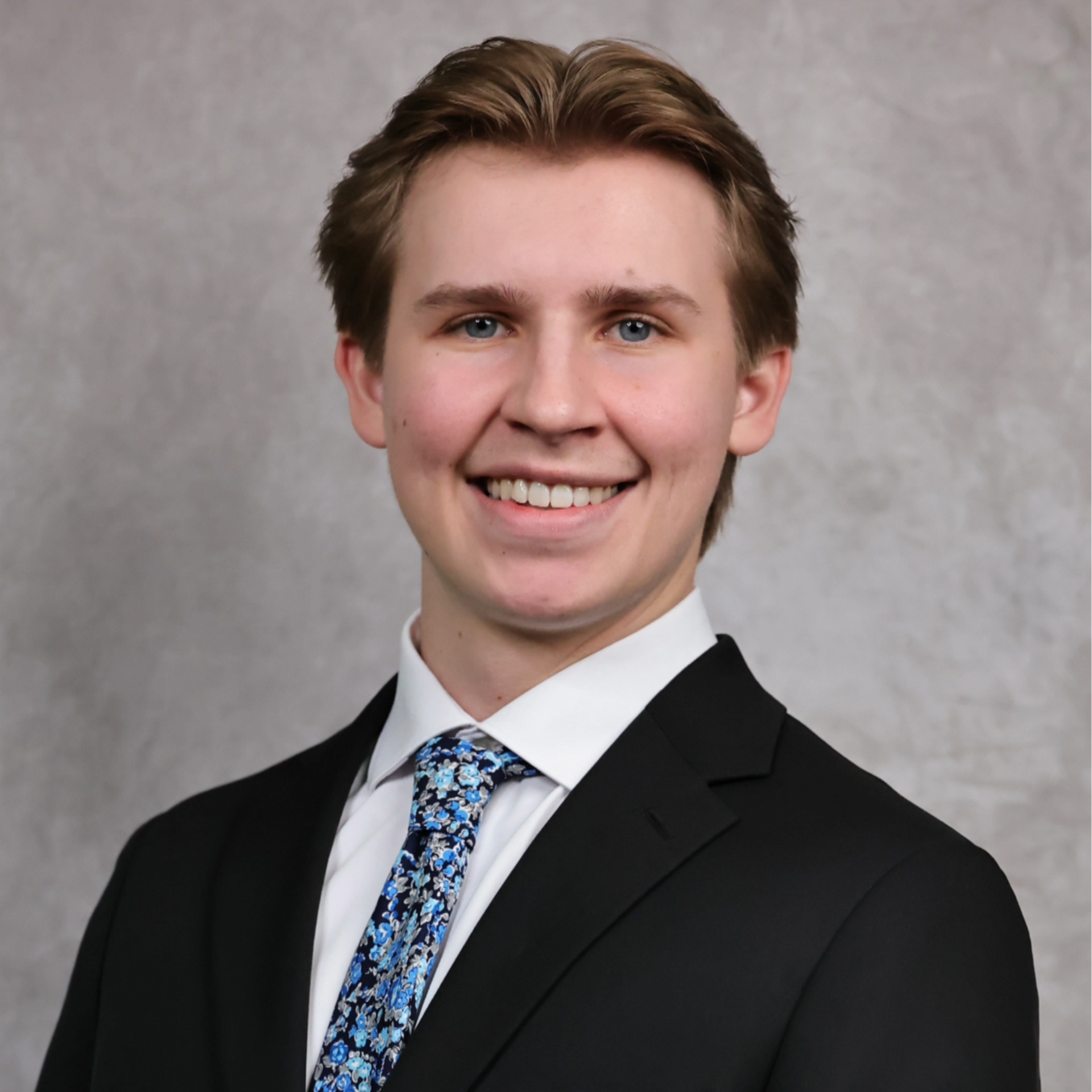 Portrait of a smiling person in a black suit, white shirt, and patterned blue tie against a gray backdrop