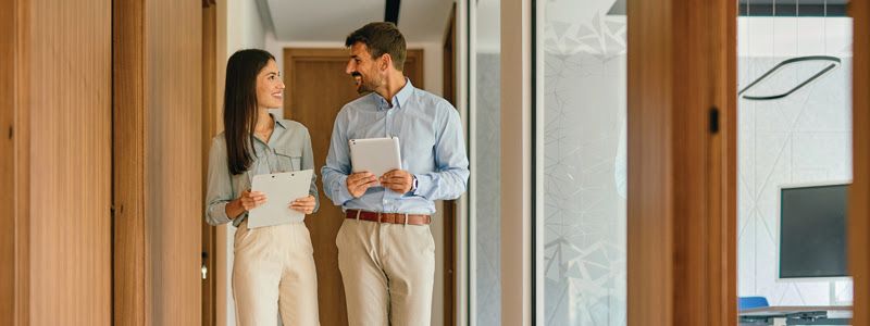 Two people standing in a doorway, talking and holding mugs and papers in a bright office hallway