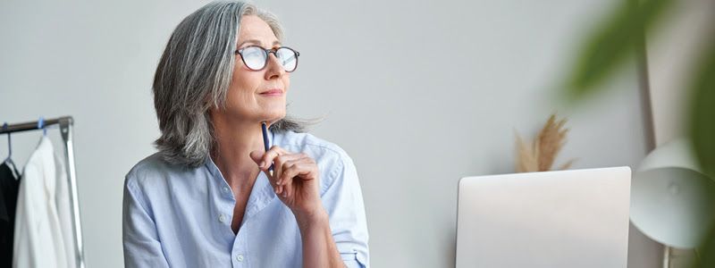 An older woman with silver hair wearing glasses and a blue shirt sits at a desk and looks out the window.