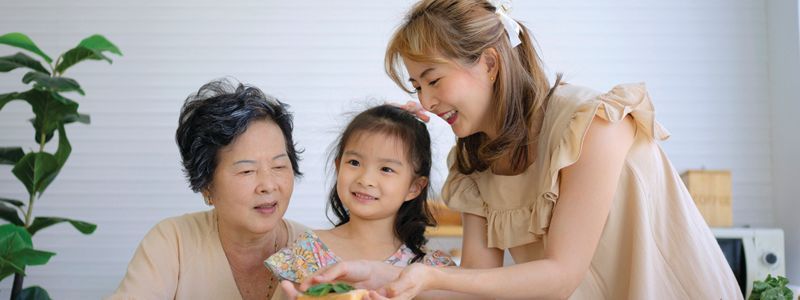 Three generations: grandmother, mother, and child looking at something, kitchen setting, light colors.