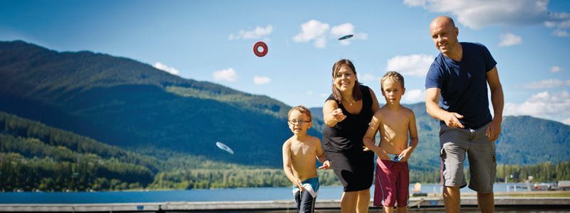 A family playing a ring toss game outdoors near a lake and mountains. Three rings are in the air, and the people are smiling.