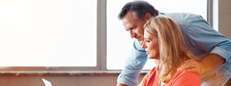 A man and woman looking at a laptop together near a window. The man has his hand on the woman's shoulder.