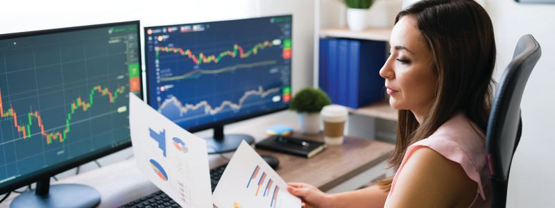 Woman analyzing stock charts on multiple monitors at a desk in a bright office