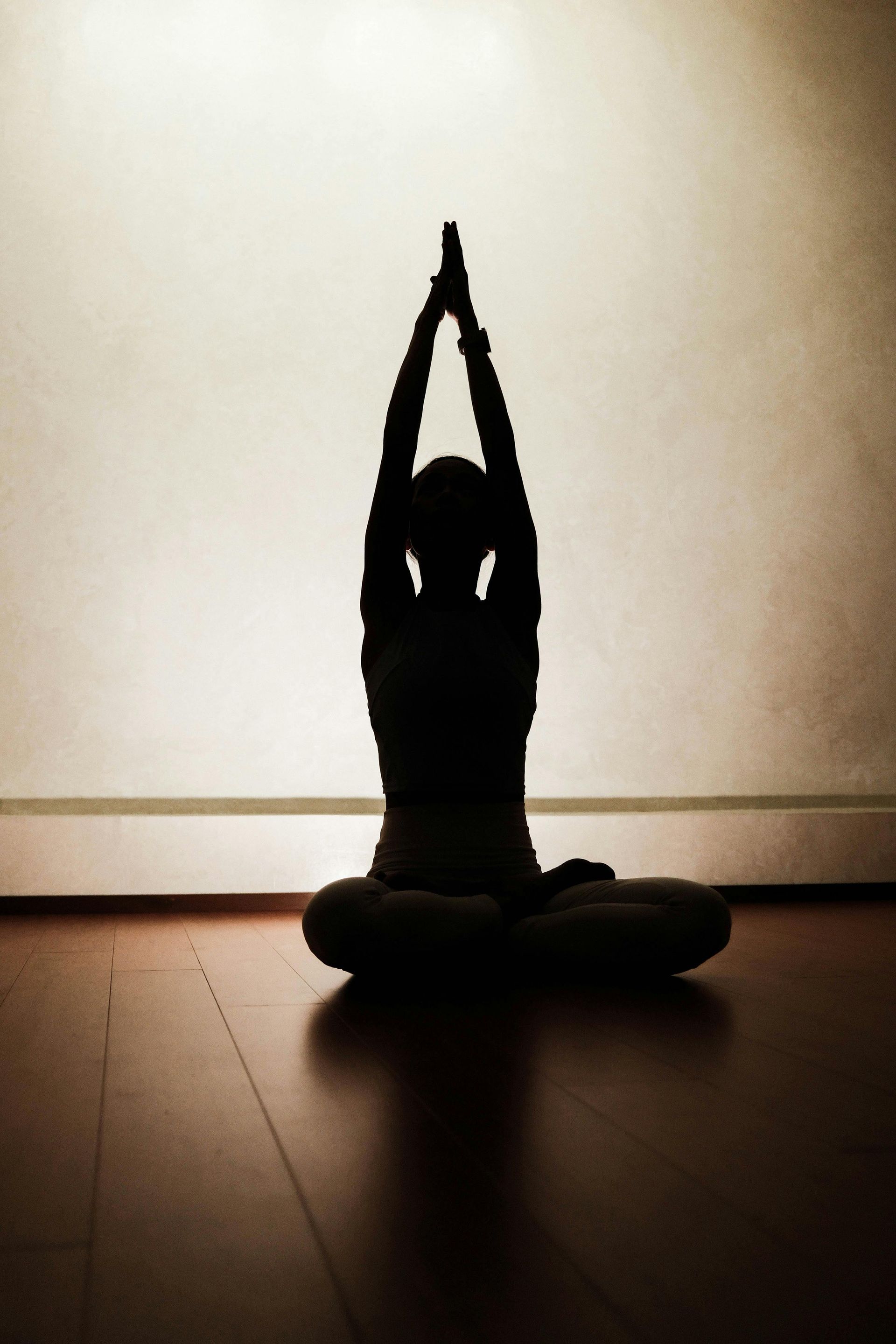 Silhouetted person in lotus pose with arms raised overhead, backlit against a wall and wooden floor
