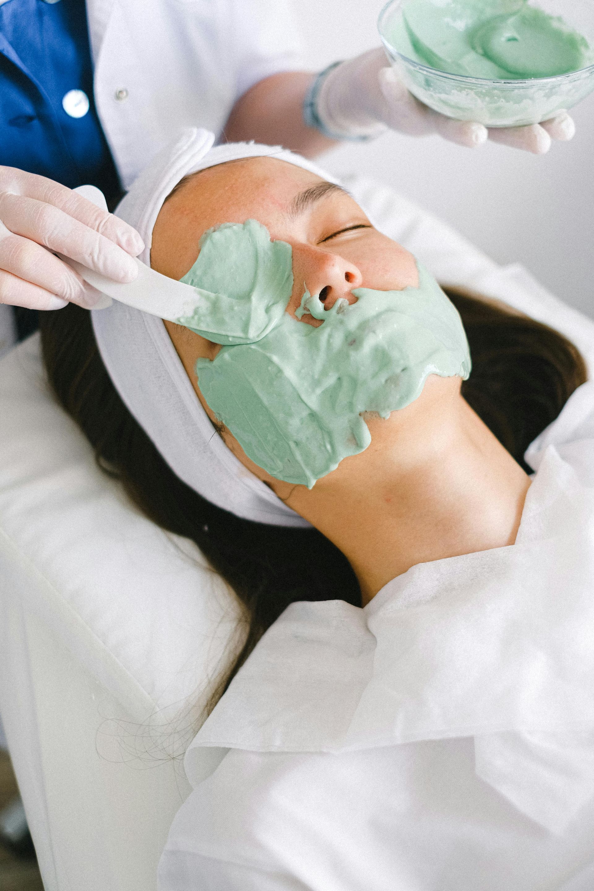 Woman receiving a mint-green facial mask treatment while lying on a spa bed