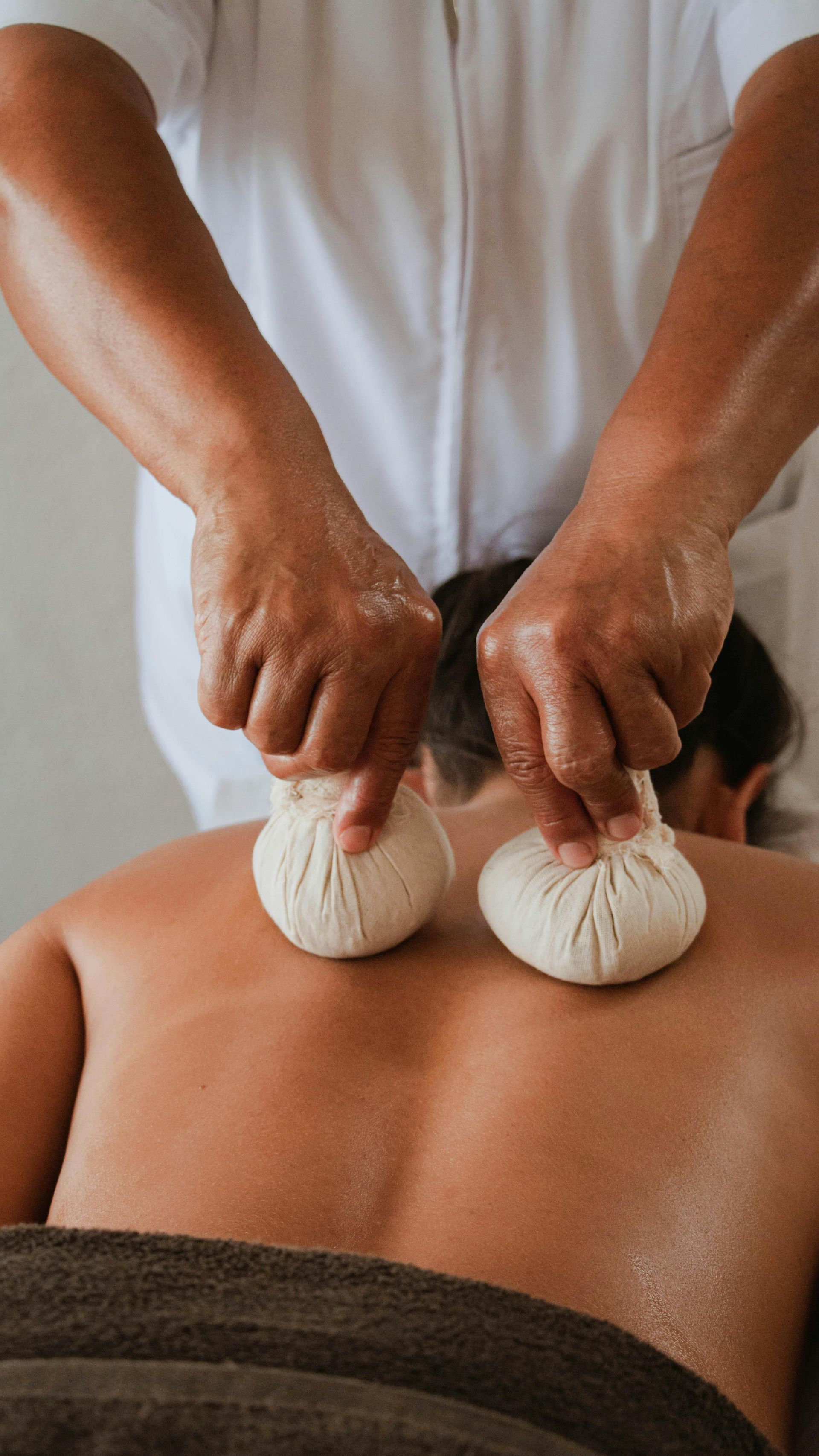 Two hands pressing herbal compresses on a person's bare back during a massage