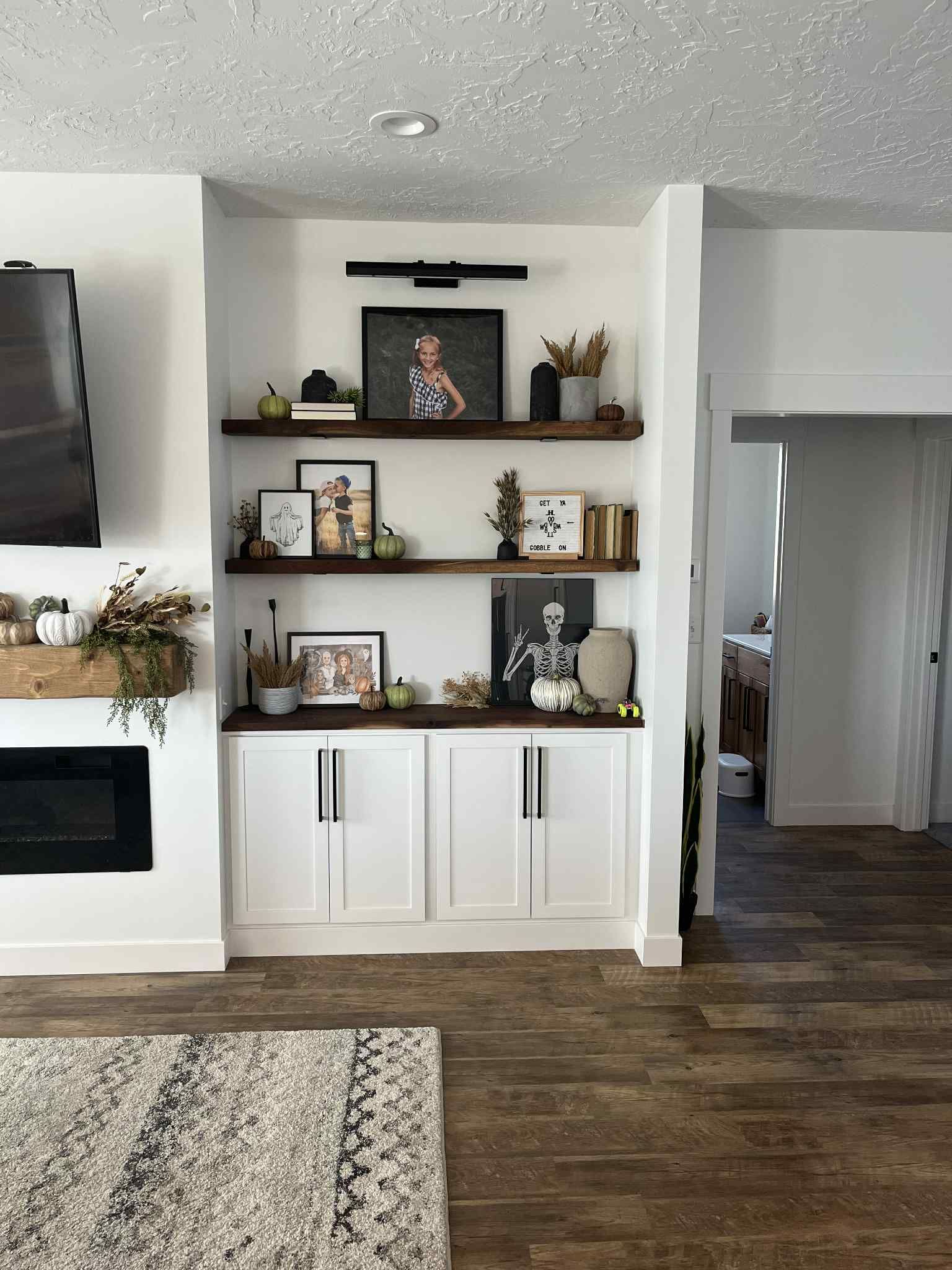 Built-in white cabinets with open shelves holding fall decor, beneath a light fixture. Wooden floor with area rug.