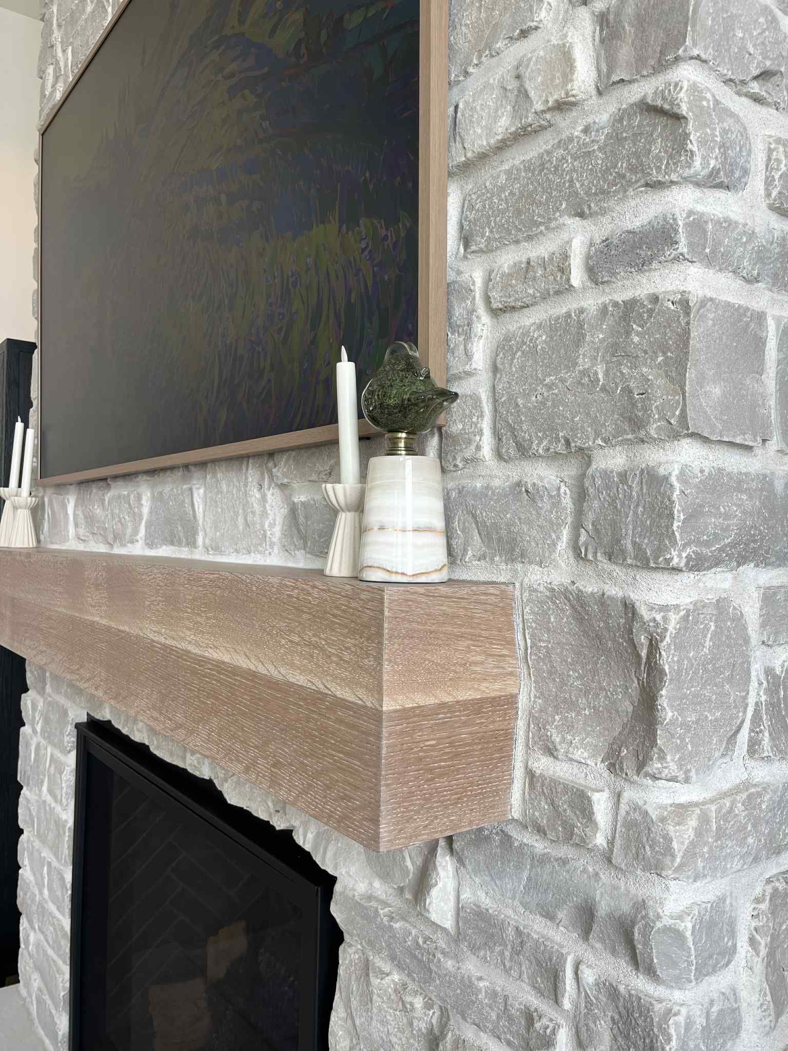 Stone fireplace with wooden mantel, holding candles and plant; a dark canvas is above.