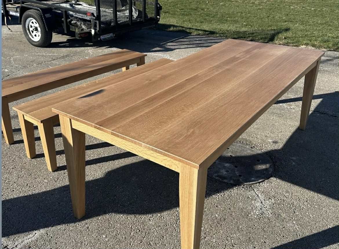 Wooden dining table and bench set outdoors, light brown color, angled legs, trailer in background.