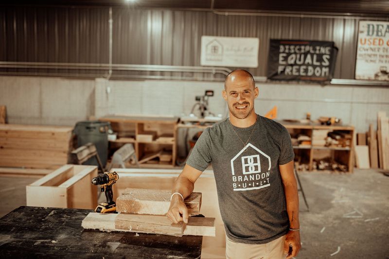 Man in workshop, smiling, leaning on table with wood and tools. Sign says 