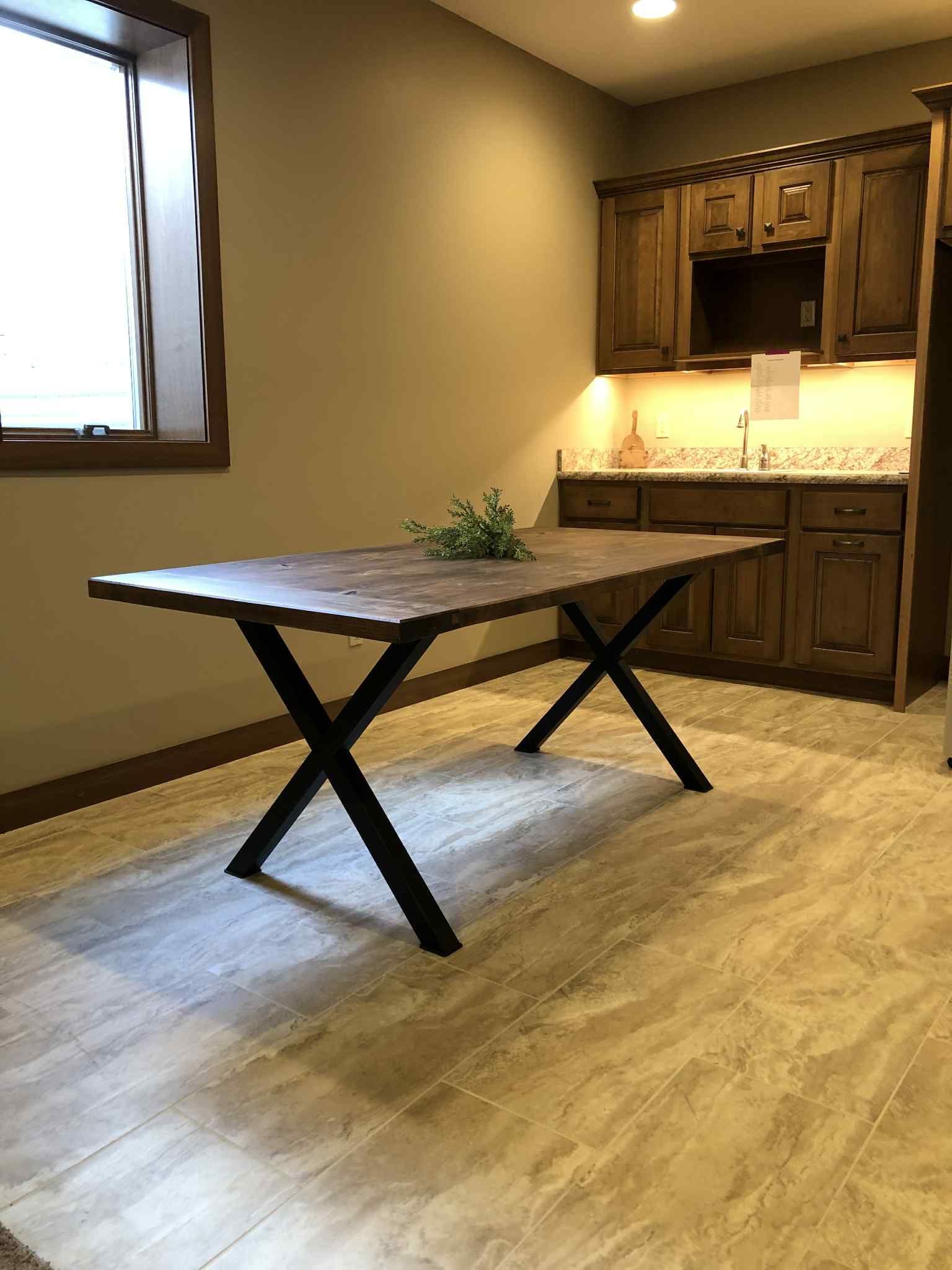 Dining room with wooden table on black cross legs; cabinets and window in background.