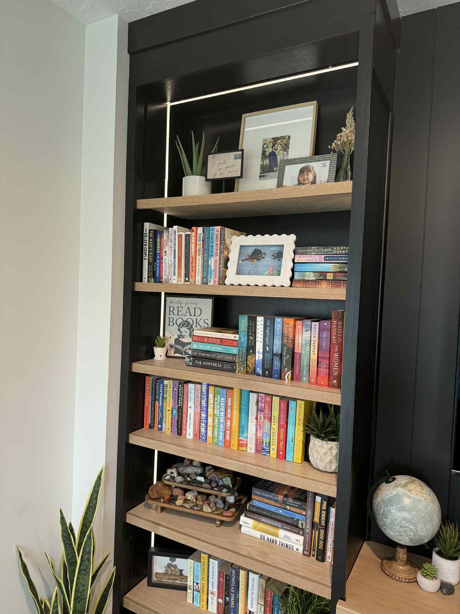 Black bookshelf with light wood shelves holding books, decor, and plants.