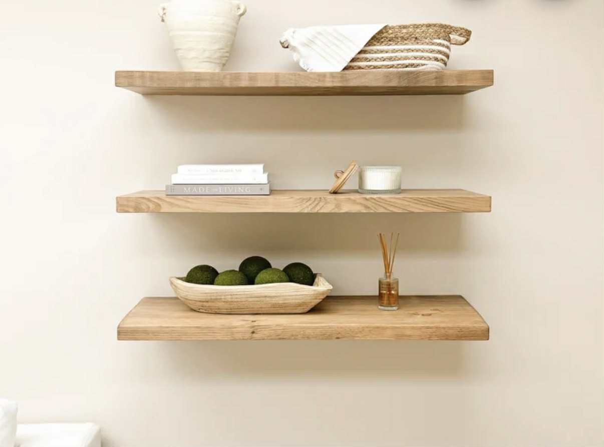 Three rustic wooden shelves on a beige wall, displaying decor: bowls, books, candles, and woven basket.