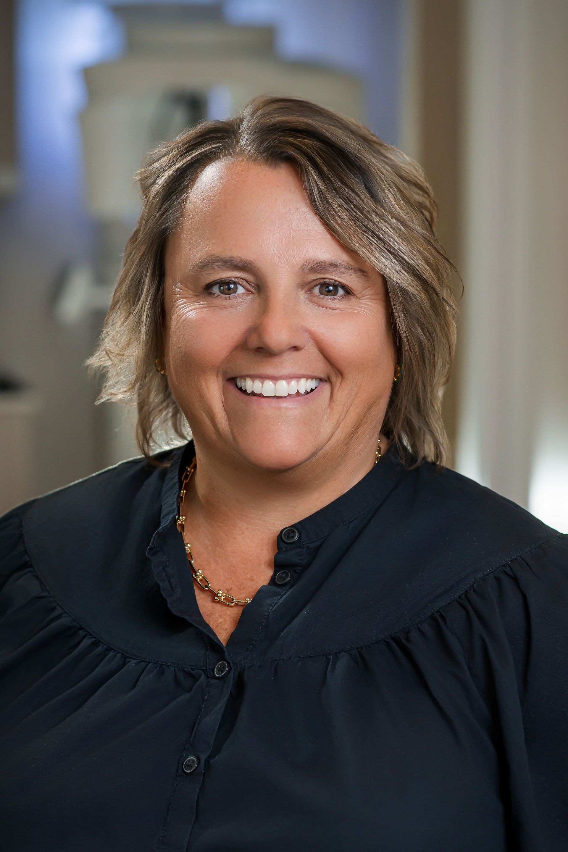 Woman in dark blouse smiles at camera, possibly in office setting.