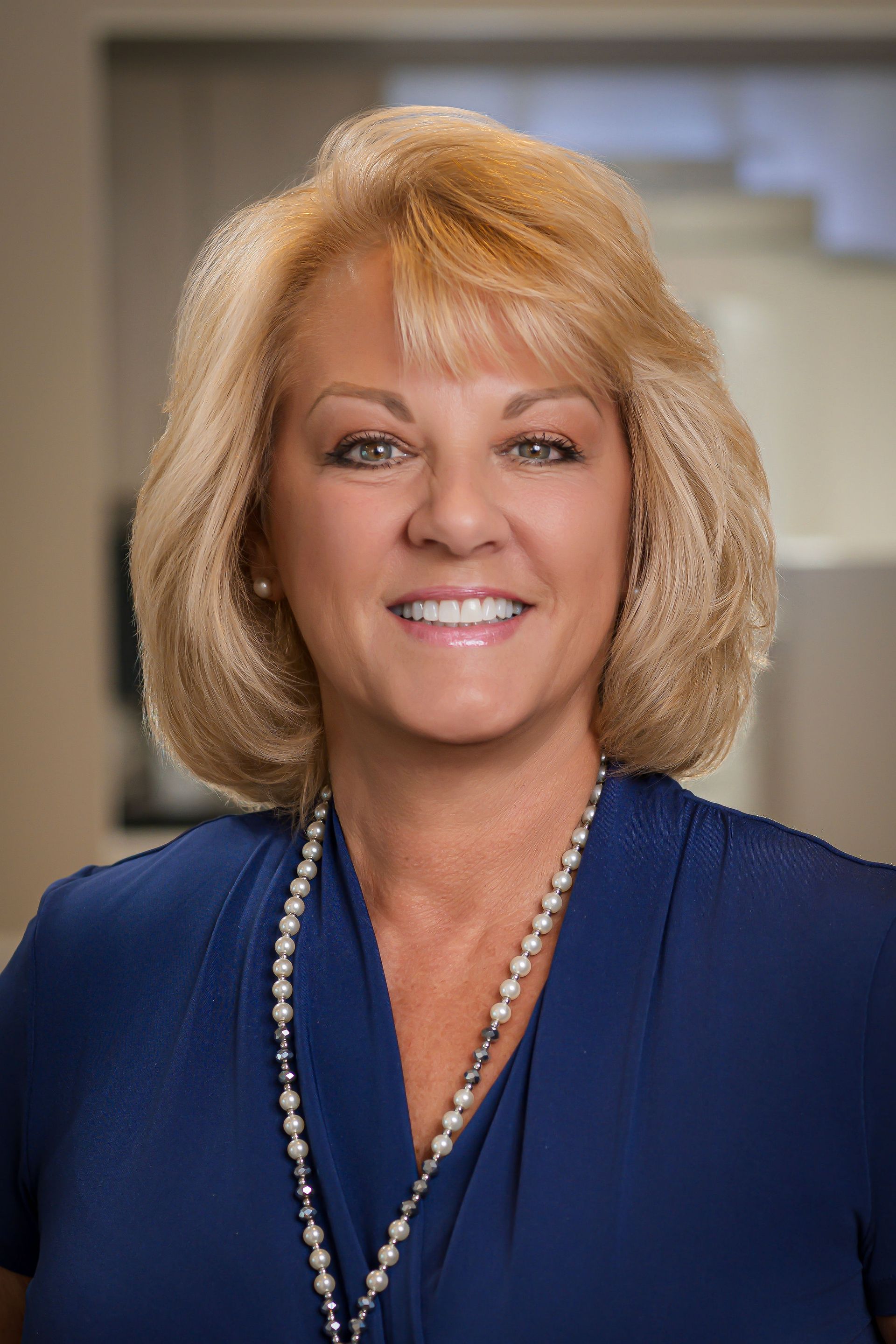 Woman with short blonde hair and a pearl necklace smiles in a blue top.