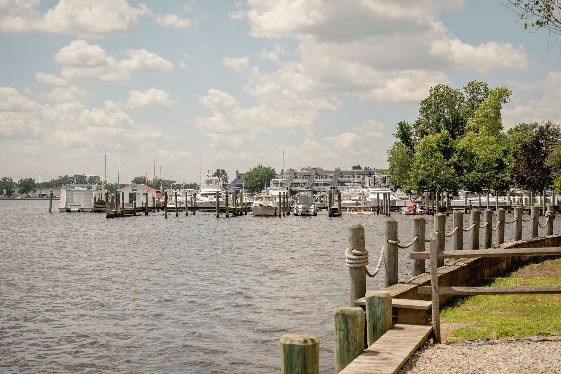 Waterfront with boats docked, cloudy sky, wooden fence in foreground, trees on the right.
