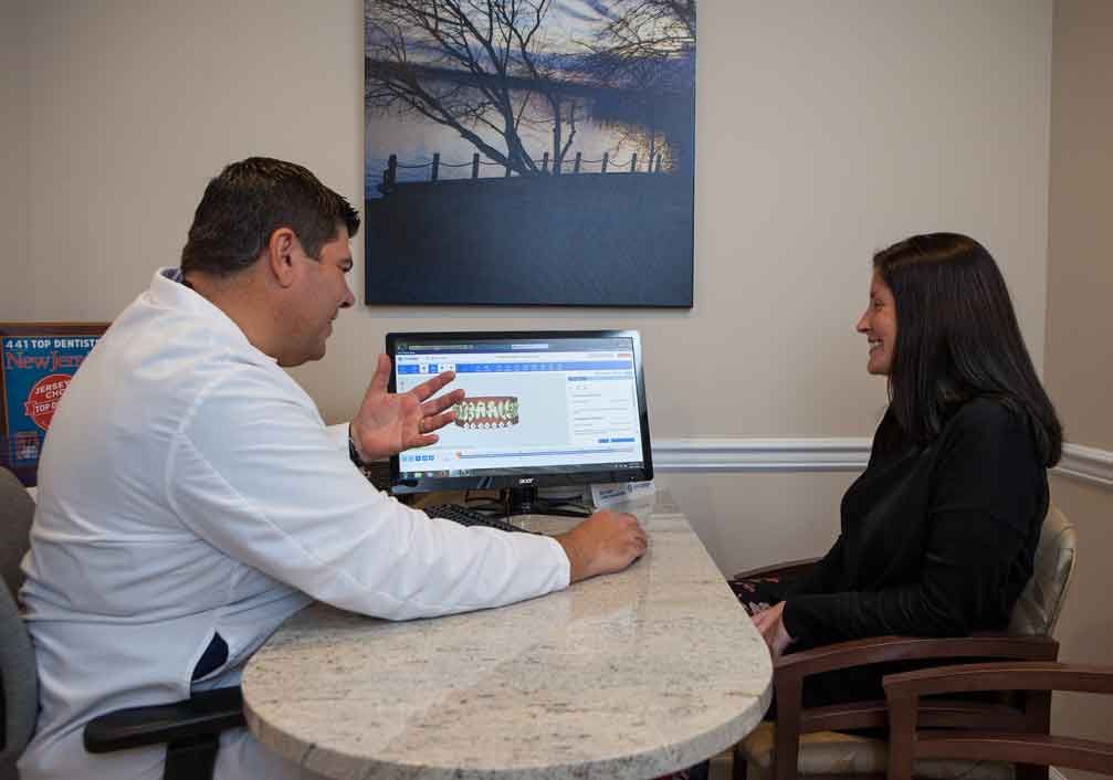 Dentist and patient at desk, reviewing x-rays on a computer monitor. Office setting.
