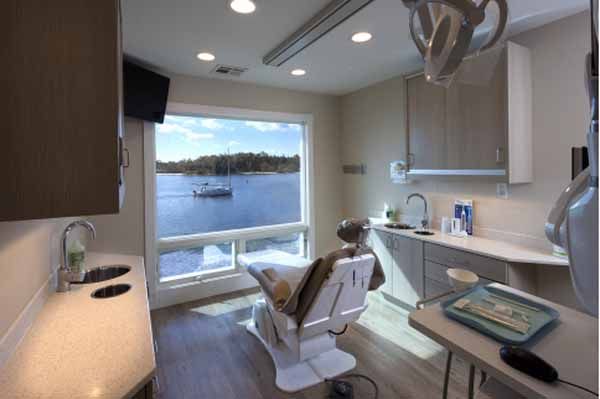 Dental office with a waterfront view. Chair faces a large window overlooking water and a boat. Cabinets and sink present.
