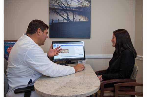 Dentist in white coat consults with a patient at a desk, looking at computer screen, soft lighting.
