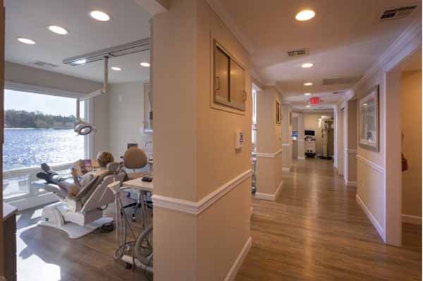 Interior view of a dental office with a patient in a chair by a window overlooking water. Corridor with columns.