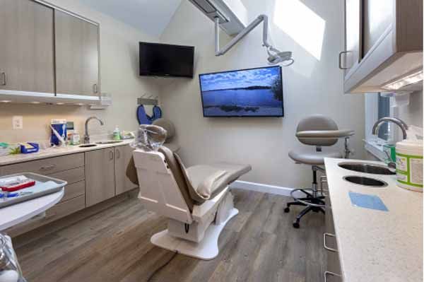 Dental operatory with a patient chair, sink, cabinets, and two wall-mounted televisions.