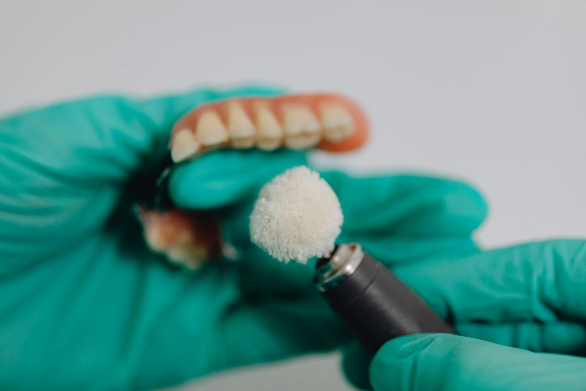 Person in green gloves polishing dentures with a rotary tool.