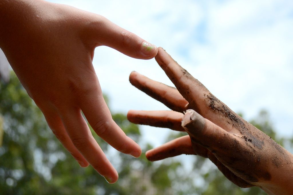 Two hands reaching toward each other; one clean, one covered in dirt, against a sky background.