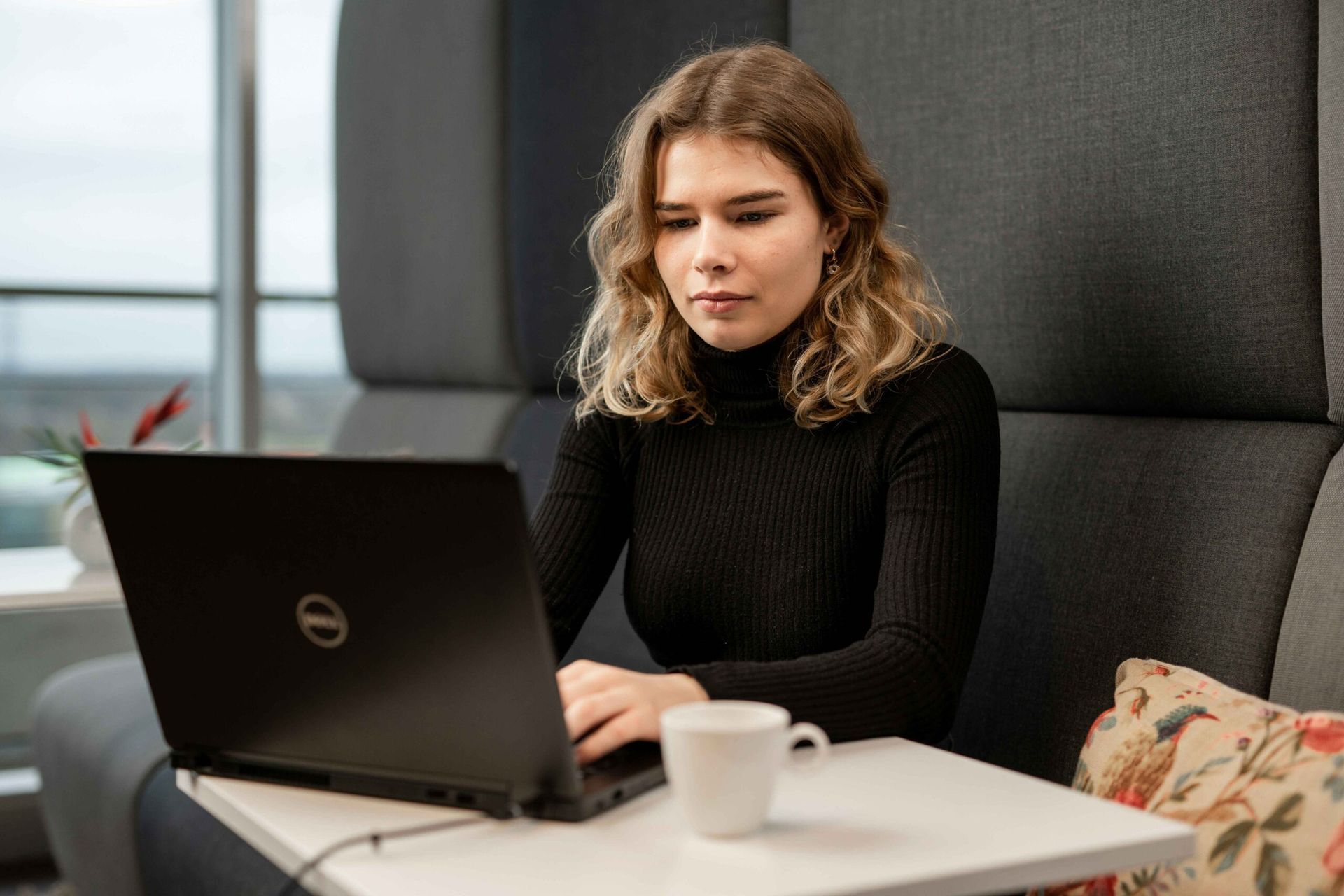 Woman in black turtleneck typing on a laptop in a modern office setting with a coffee cup nearby.