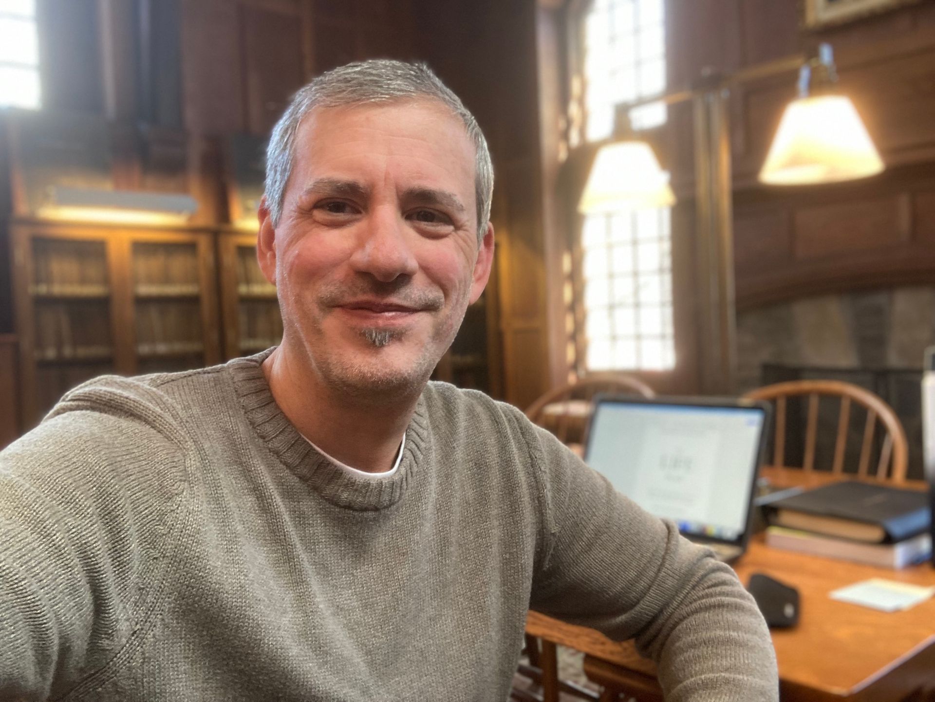 Man smiling at a desk in a wood-paneled library, with a computer and lamp nearby.