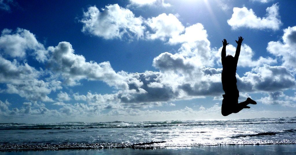Silhouette of person jumping for joy on beach with blue sky and clouds.