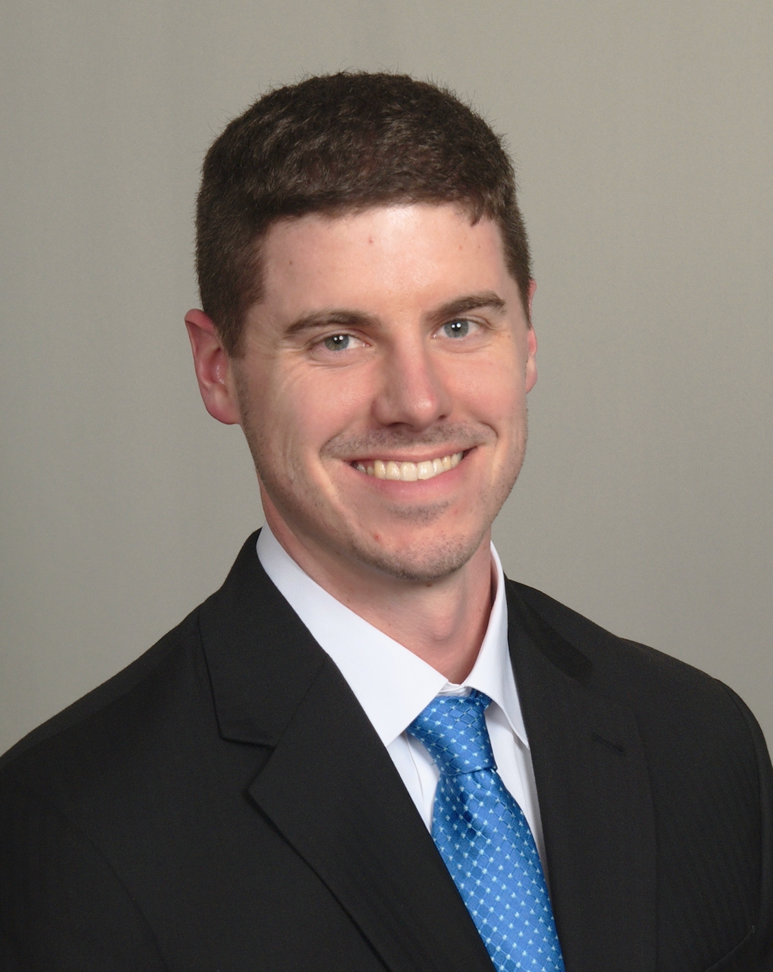 Smiling man in a black suit, white shirt, and blue tie against a gray background