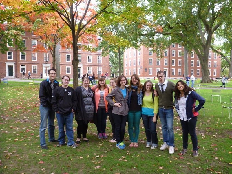 Group of diverse students pose on a green lawn in front of brick buildings with fall foliage.
