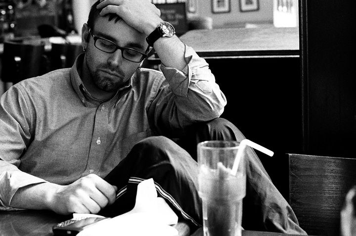 Man with glasses slumped at a table, hand on head. He has a drink with a straw.