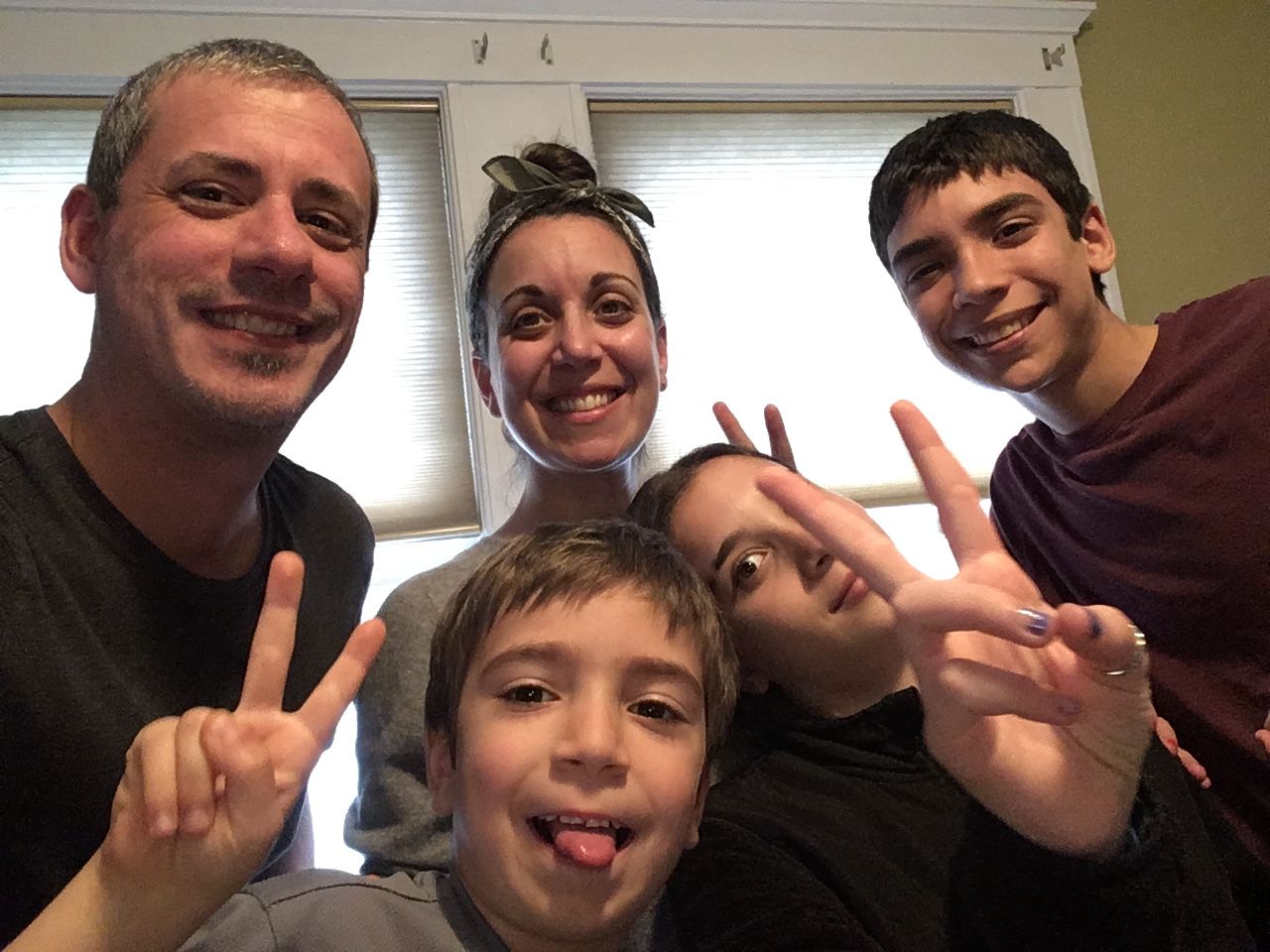 Five people smiling and posing indoors, several making peace signs in front of a window.