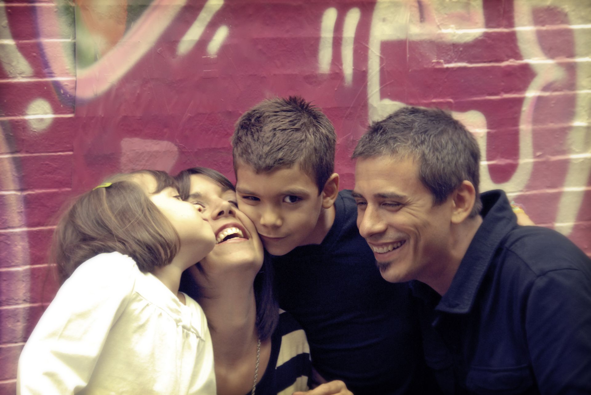 Two children and a man smiling and embracing in front of a colorful graffiti wall