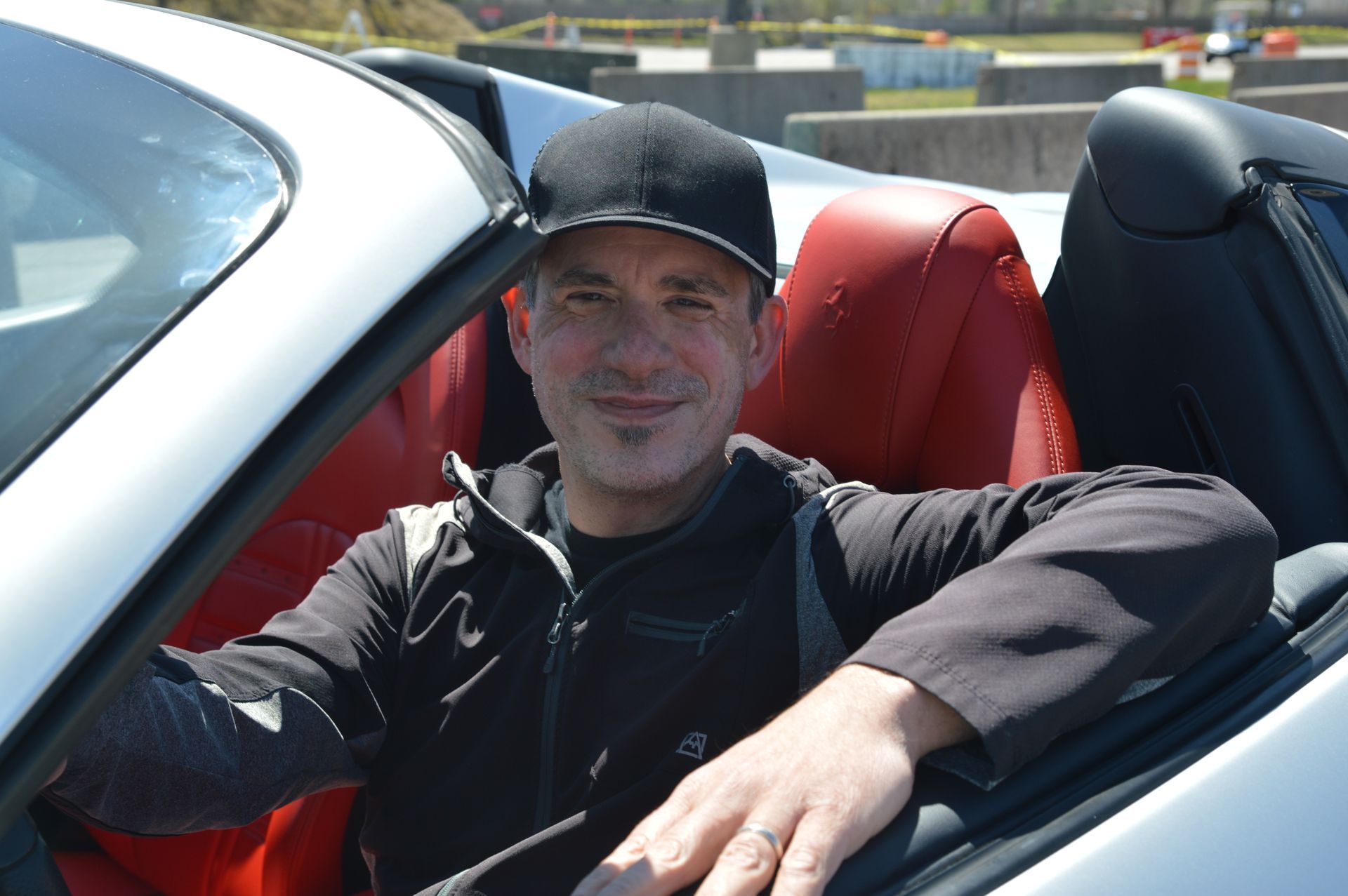 Man sitting in a red convertible, wearing a black cap and jacket, smiling in bright sunlight