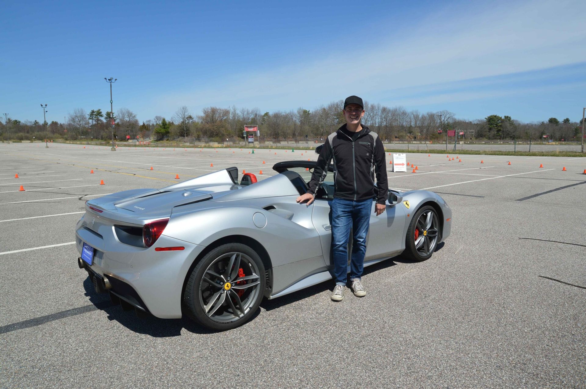 Man in black jacket and jeans stands next to a silver Ferrari convertible on a sunny asphalt track.