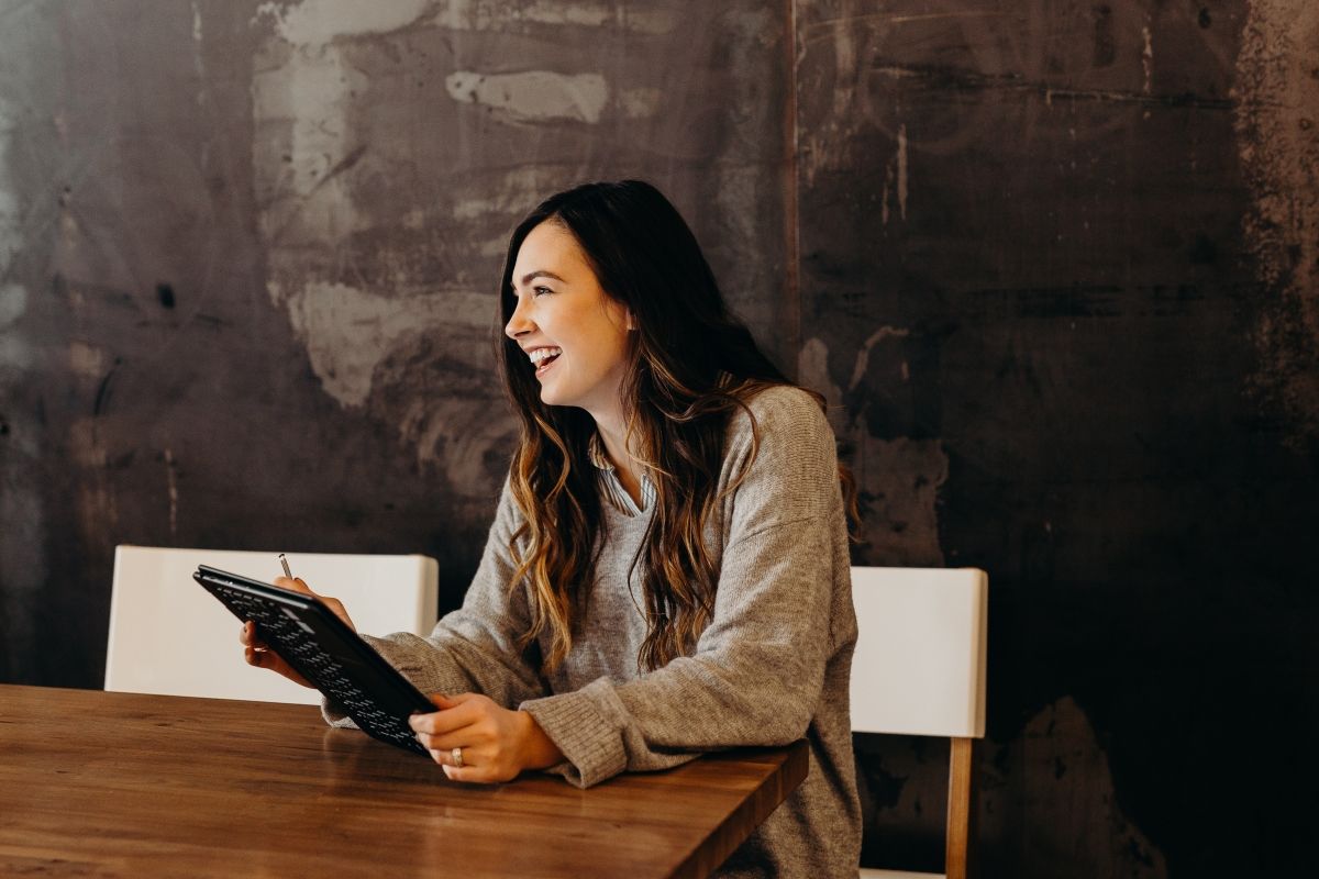 Woman with long brown hair laughing, sitting at a table, holding a tablet. Neutral background.