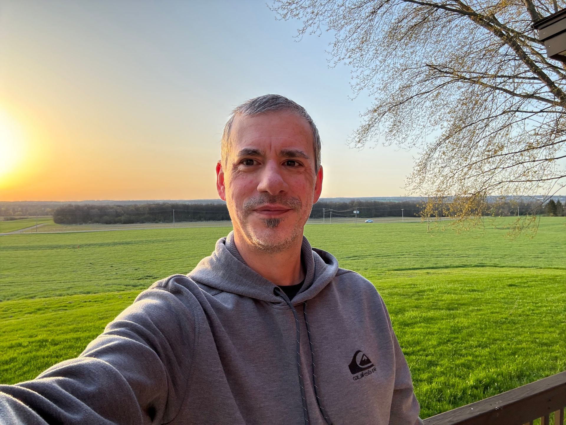 Selfie of a person in a gray hoodie standing in a green field at sunset