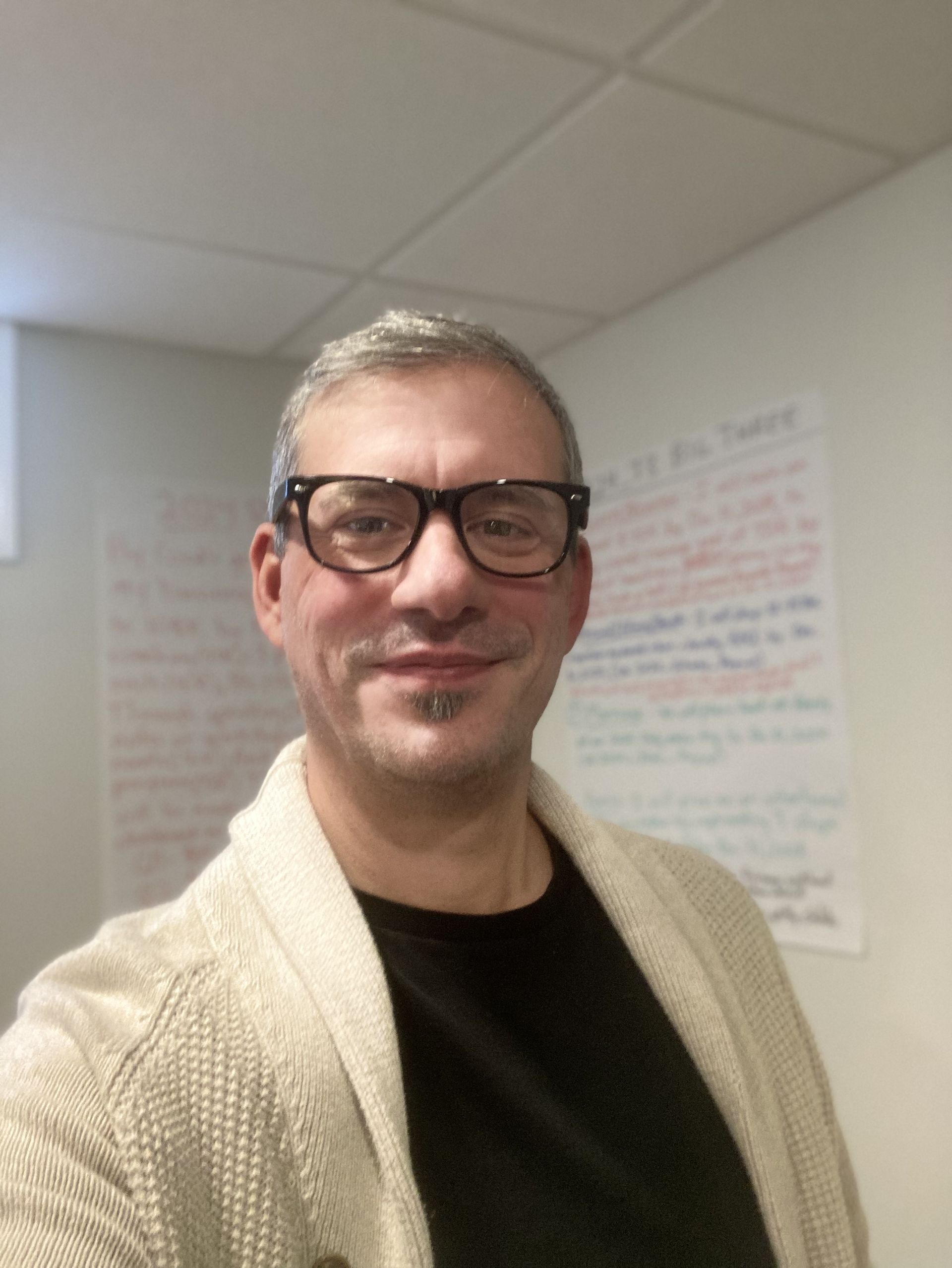 Man with glasses smiles, wearing a beige cardigan over a black shirt, in front of a white wall with writing.