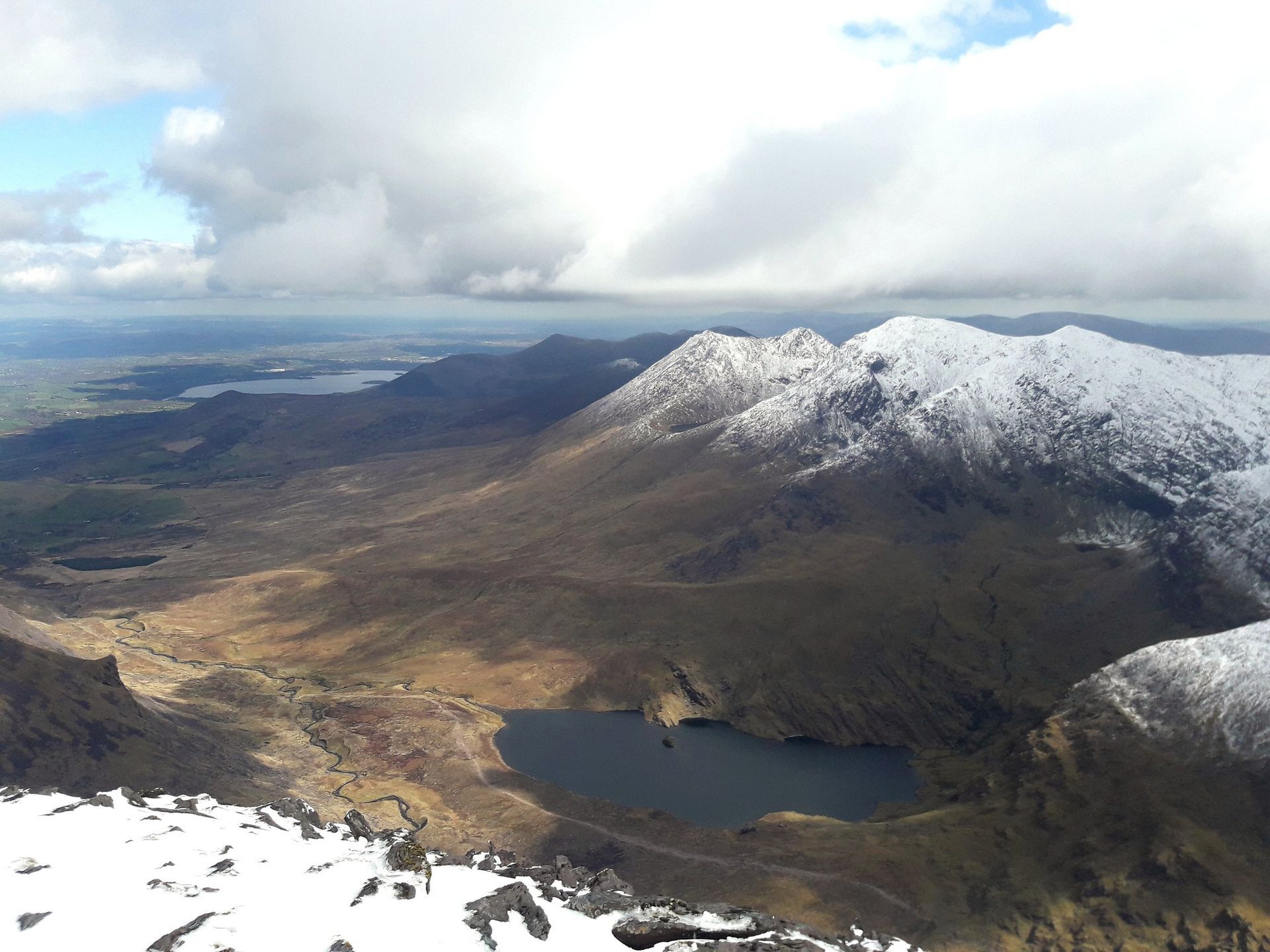Snowy mountain peaks overlooking a lake and valley under a cloudy sky.
