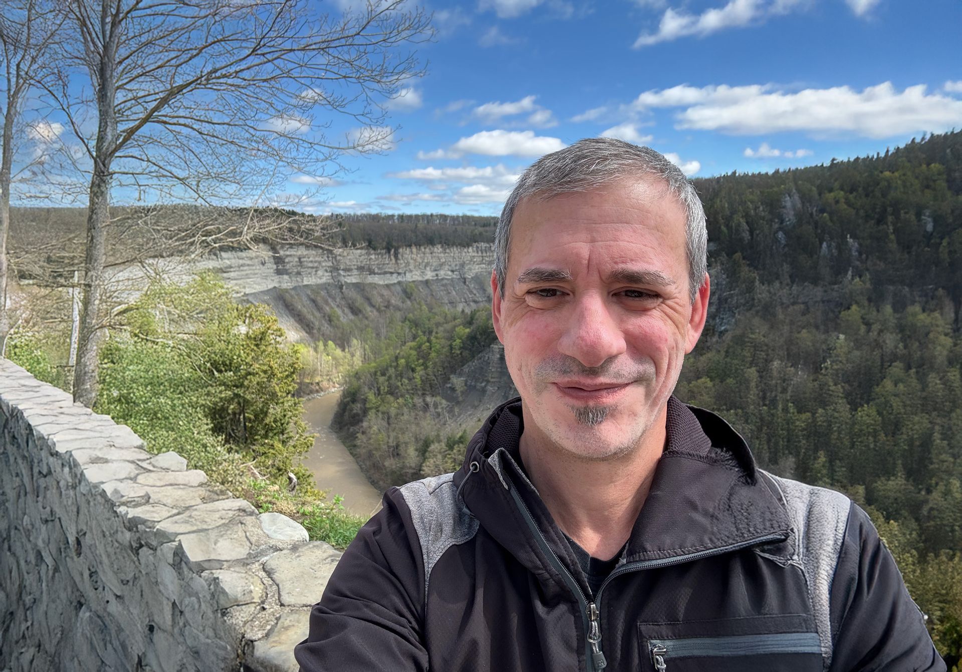 Man taking a selfie by a stone wall with a canyon and forested hills under a blue sky