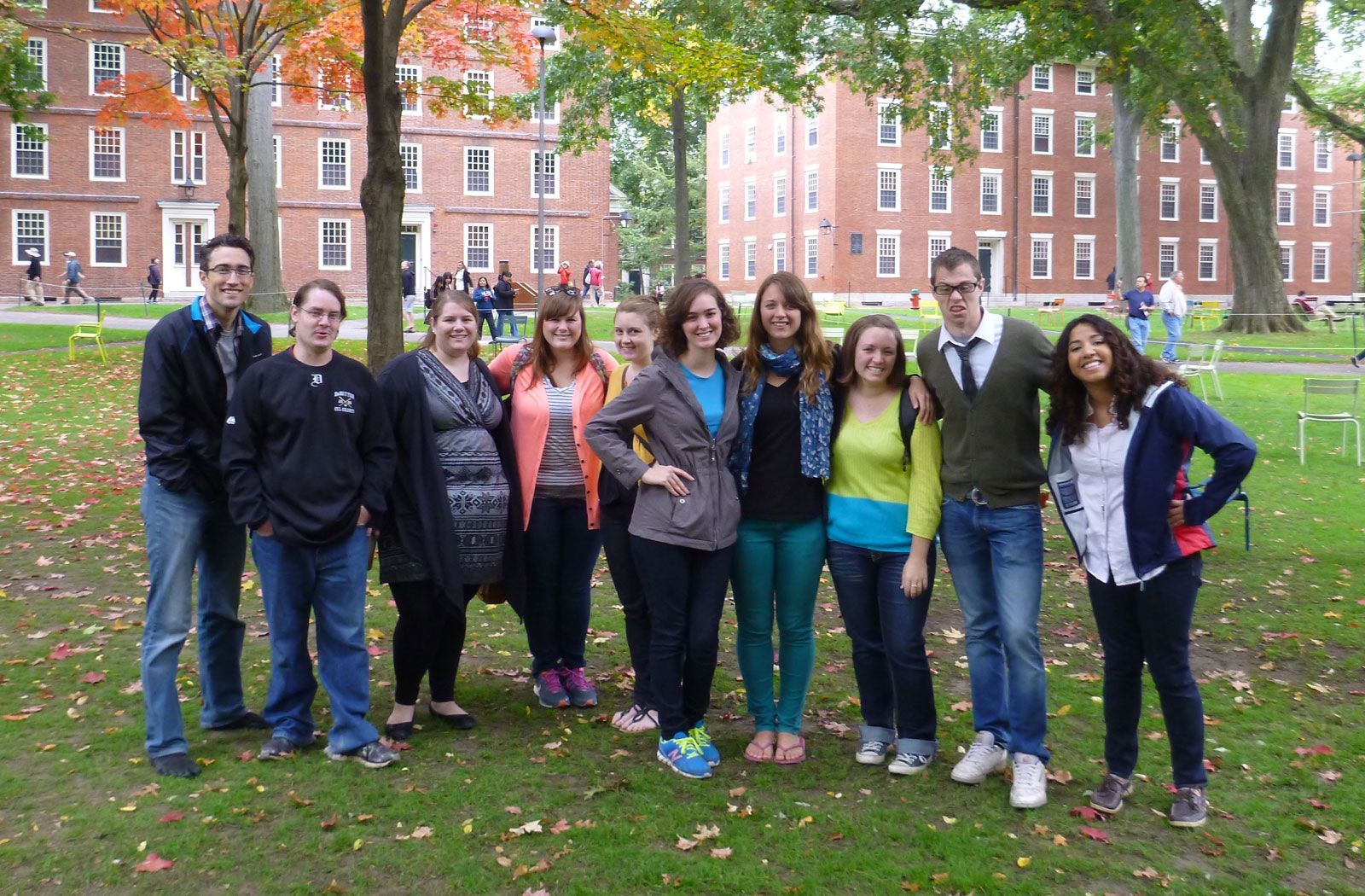 Group of students posing on a green lawn in front of brick buildings. Autumn foliage.