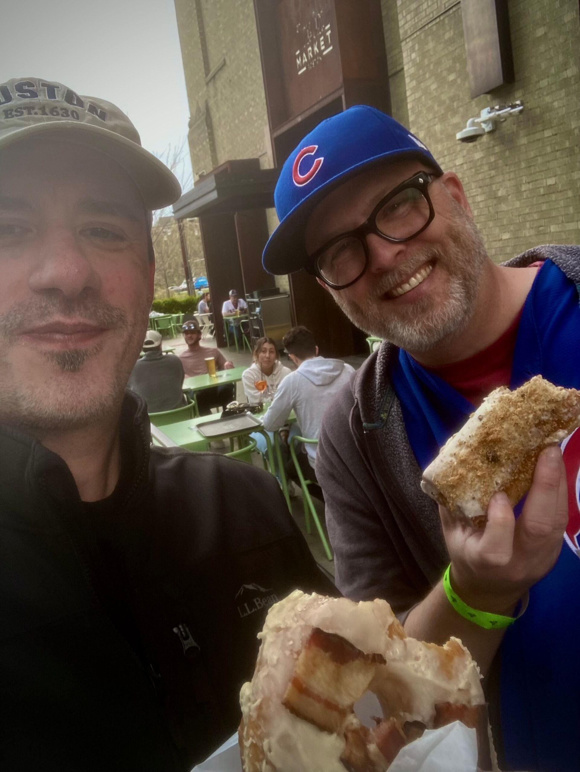 Two men smiling, eating sandwiches outdoors. One wears a Cubs hat, the other a cap.